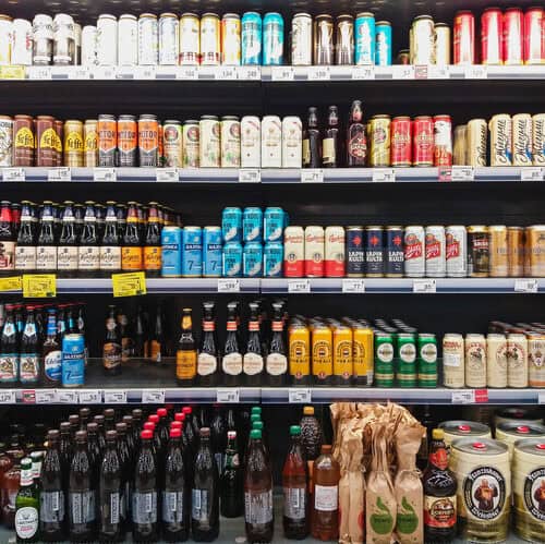 Grocery store beer aisle with rows of beer bottles and cans on shelves.”] ,