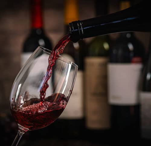Red wine being poured into a tilted wine glass with blurred bottles in the background, creating a moody still-life scene.